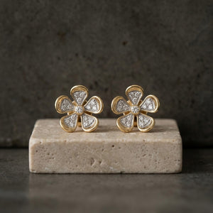 A pair of gold flower-shaped stud earrings with clear pavé-set natural diamonds stones, displayed on a rectangular stone block against a dark textured background.
