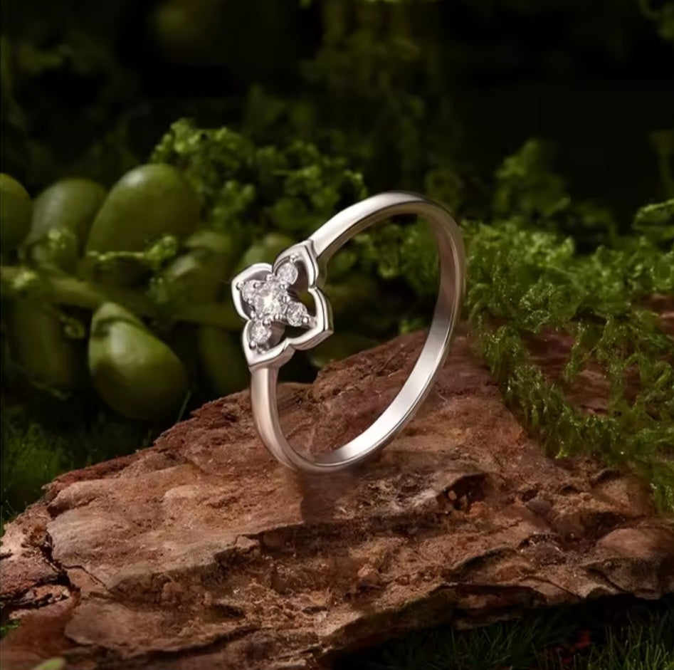 A silver ring with a four-petal flower setting and clear gemstones, resting on a textured brown log with green moss and leaves in the background.