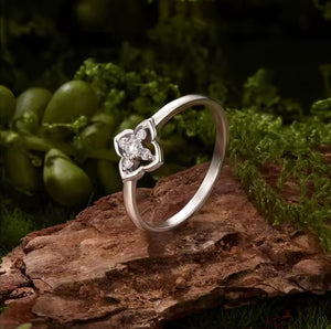 A silver ring with a four-petal flower setting and clear gemstones, resting on a textured brown log with green moss and leaves in the background.