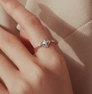 A silver ring with a four-petal flower setting worn on a person's ring finger, shown against a soft beige ribbed fabric background.