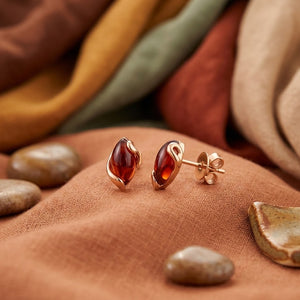 A pair of rose gold stud earrings with polished amber-colored marquise-cut stones, displayed on a textured rust-brown fabric alongside smooth stones and soft draped textiles in the background.