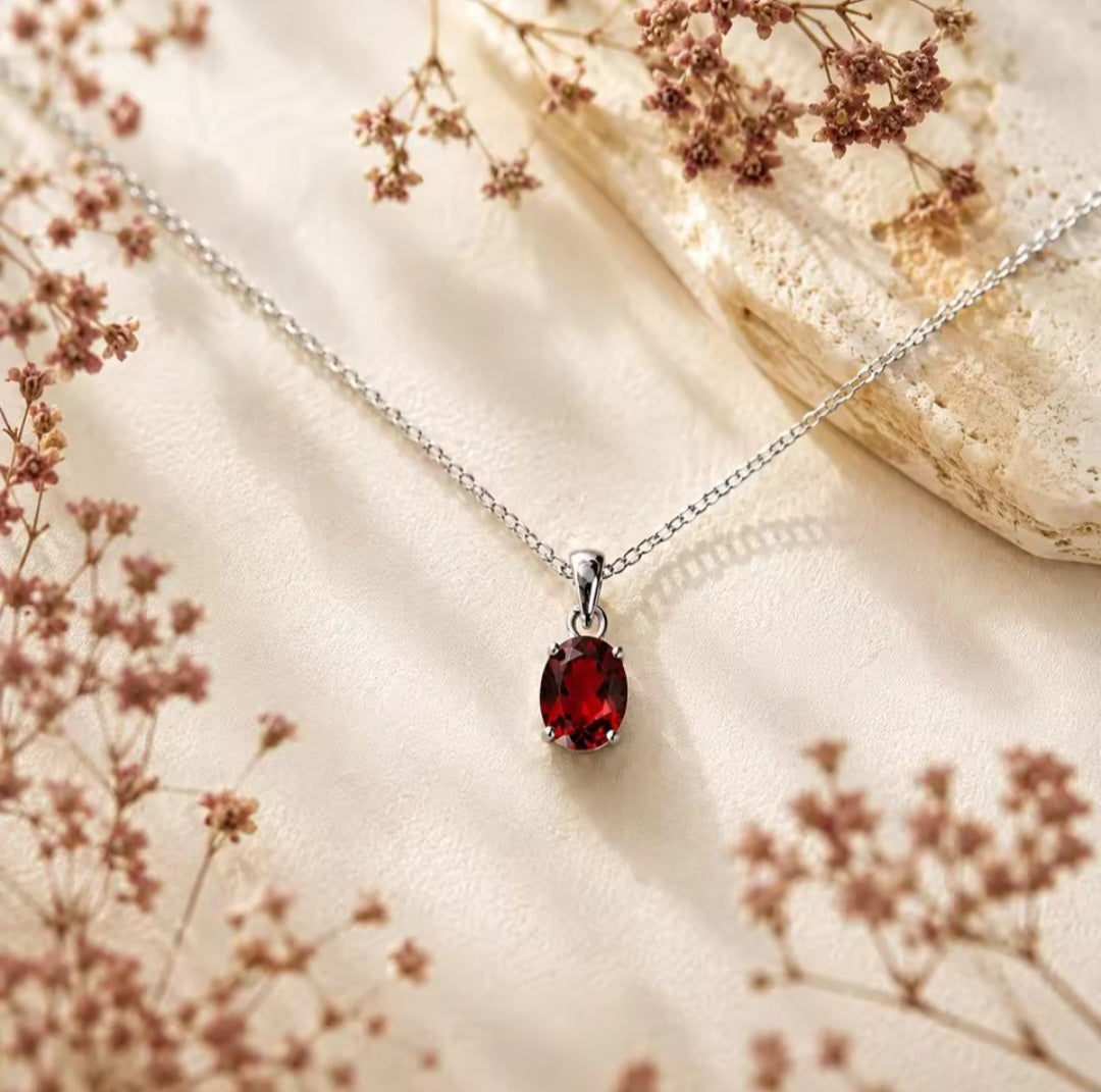 A silver chain necklace with an oval red natural garnet gemstone pendant, displayed on a textured cream fabric with dried floral elements.