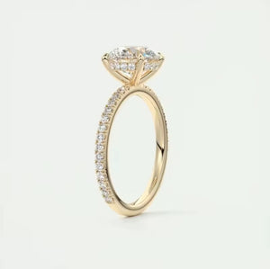 A yellow gold ring featuring a large round cut clear moissanite gemstone in a four-prong setting, with a pave band adorned with small round-cut stones, shown on a light grey background.