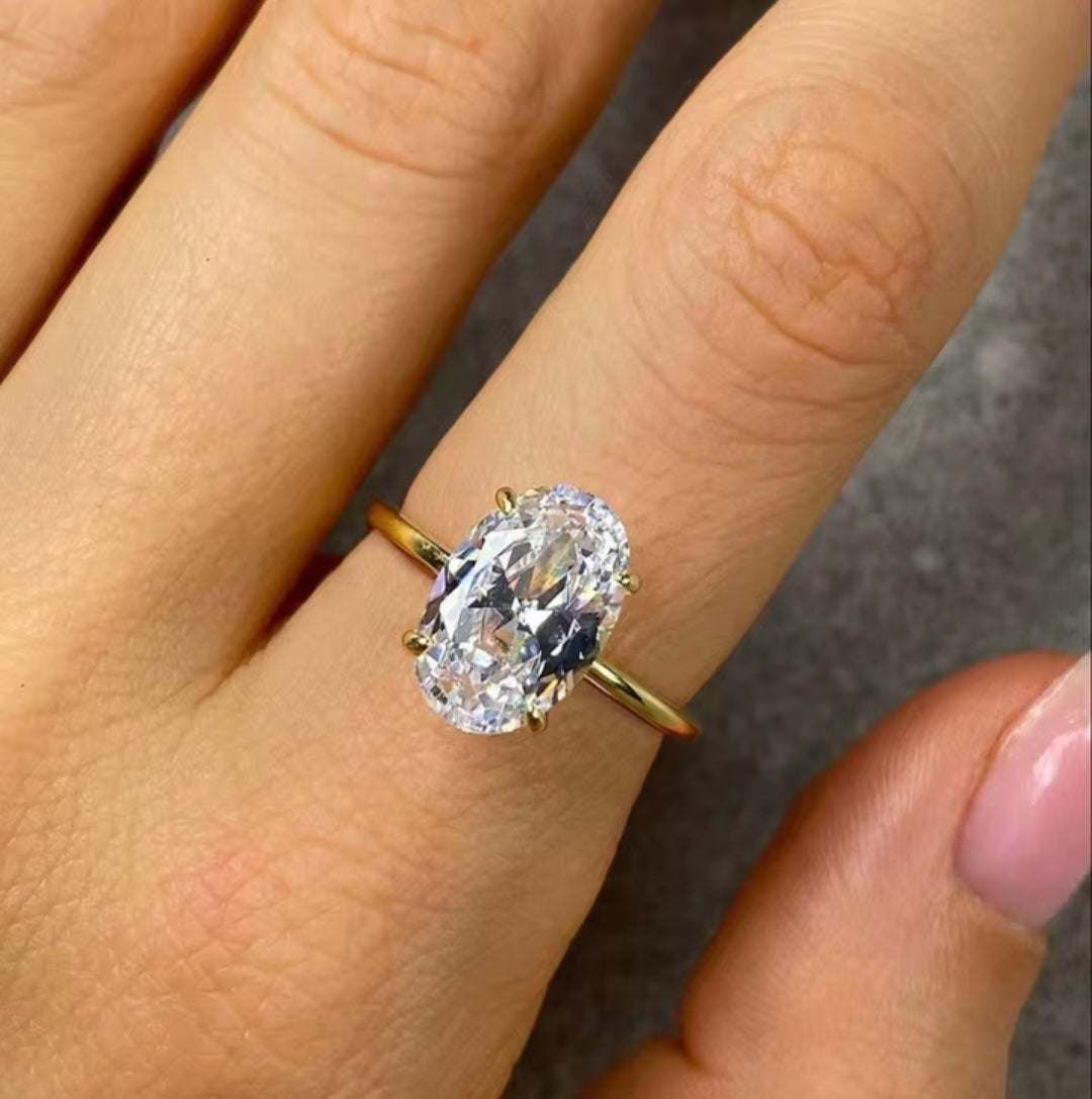An oval-cut clear gemstone ring set in a yellow gold band, shown worn on a person's finger.
