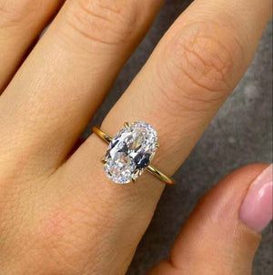An oval-cut clear gemstone ring set in a yellow gold band, shown worn on a person's finger.
