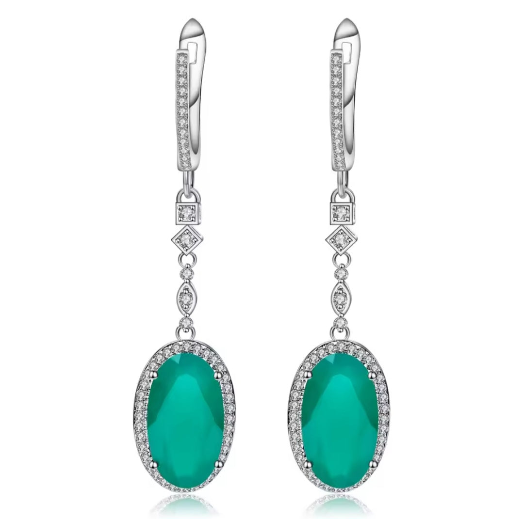 A pair of silver drop earrings featuring oval emerald-cut green gemstones surrounded by small clear stones, with leverback hooks and diamond-shaped accents, shown on a white background.