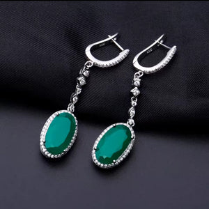 A pair of silver drop earrings with oval green gemstones and small clear stones, displayed on a dark textured fabric background.