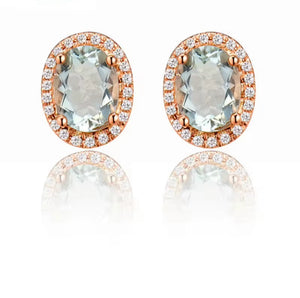 A pair of oval stud earrings featuring large pale blue-green center stones surrounded by a halo of small clear stones in rose gold settings, shown on a white background with a soft reflection below.