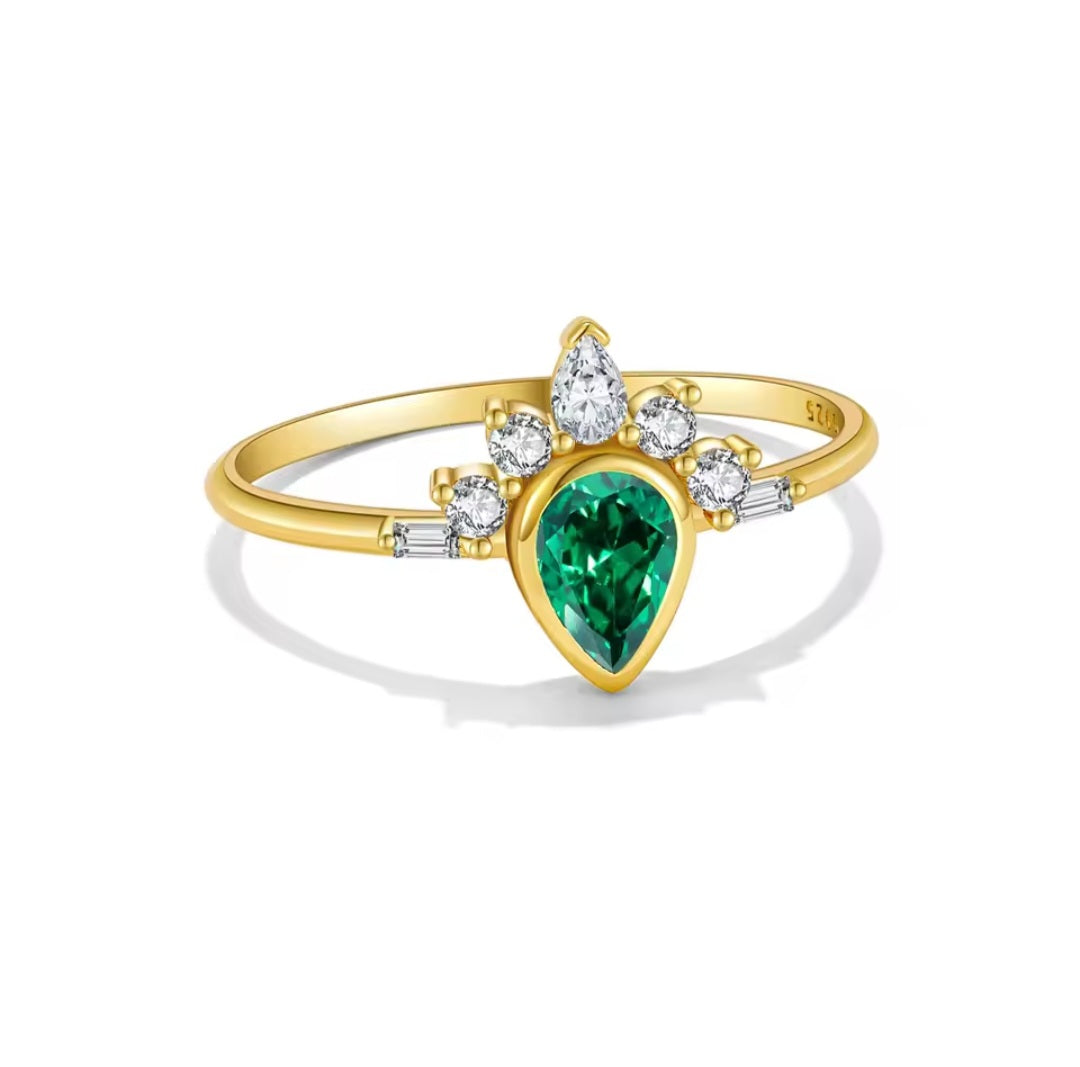 A yellow gold ring featuring a pear-cut green emerald center stone, surrounded by small round clear stones and baguette accents on a white background.