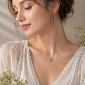 A woman wearing a delicate silver necklace with a small green teardrop green peridot pendant, dressed in a white V-neck blouse against a softly lit neutral background.