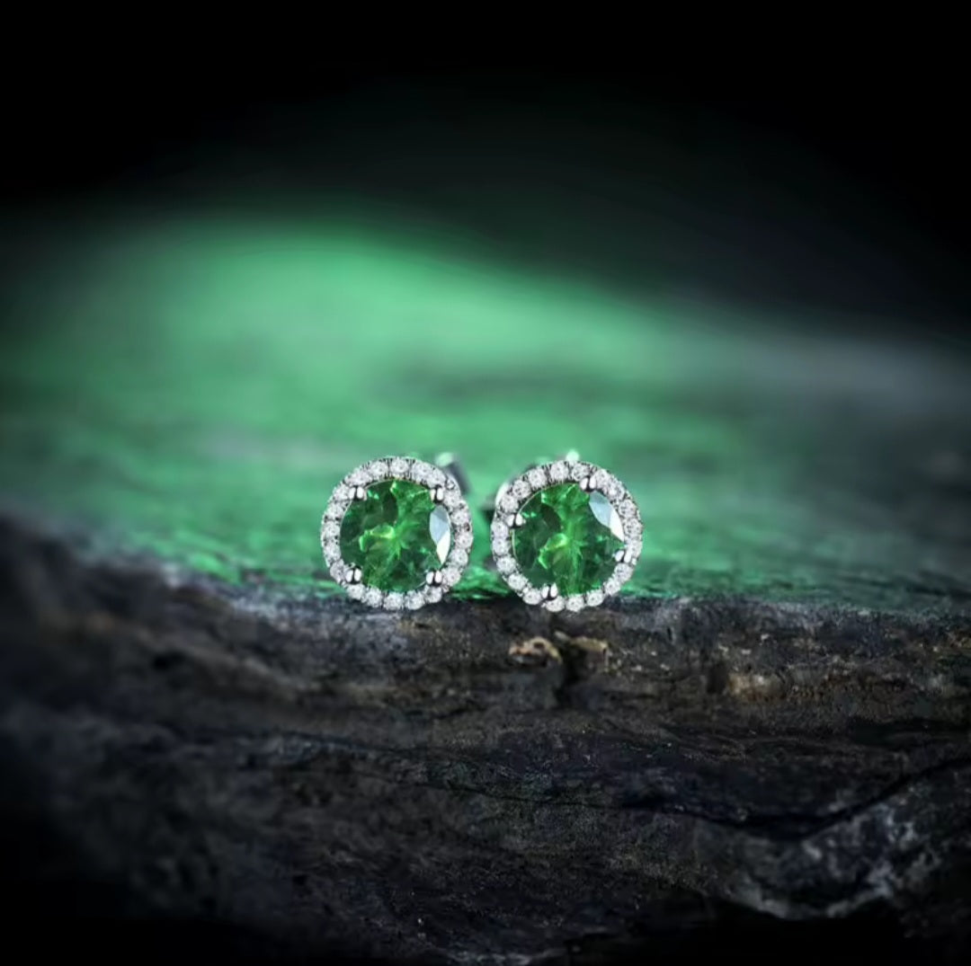 A pair of round green tsavorite gemstone stud earrings with white gold and diamond halos, resting on a dark textured surface with a glowing green background.