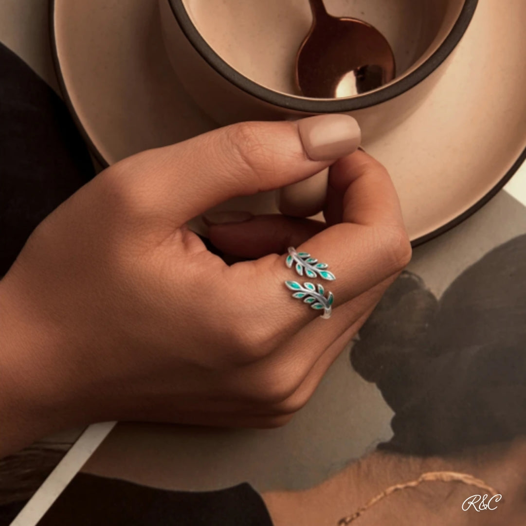 A person's hand displaying a green and silver solid silver ring with an intricate leaf pattern design, placed on a finger next to a cup and a photograph.