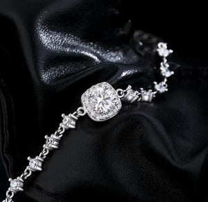 A silver chain bracelet featuring a cushion-cut clear gemstone surrounded by smaller stones, displayed on a draped black fabric background.