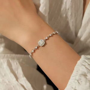 A delicate silver bracelet with a central round clear gemstone and small sparkling  moissanite stones along the band, shown worn on a person's wrist against a soft white textured garment background.