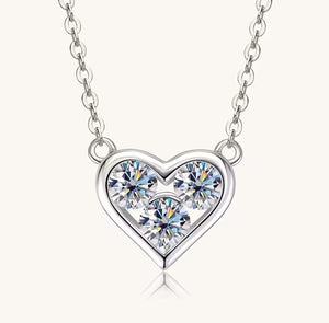 A silver heart pendant necklace featuring three sparkling clear gemstones, suspended from a delicate silver link chain against a light cream background.