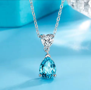 A silver pendant necklace featuring a heart-shaped clear stone and a teardrop-shaped light blue gemstone, displayed on a bright blue surface.