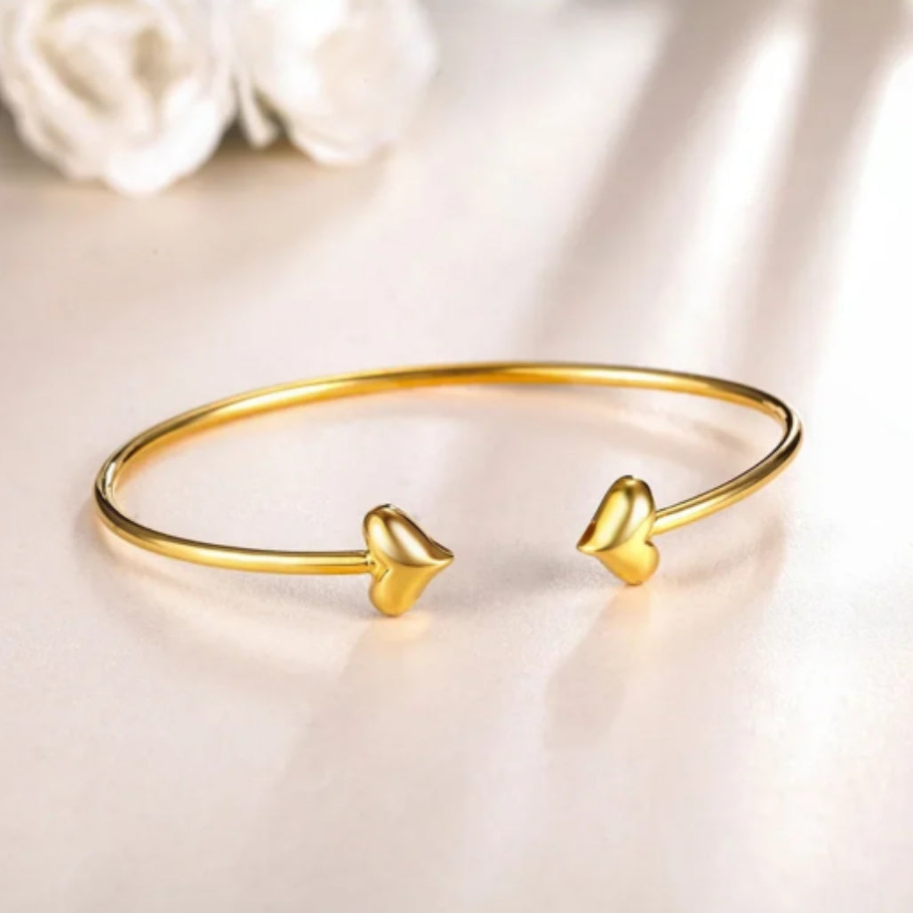 An 18K yellow gold bracelet with a delicate heart design at each end, displayed on a reflective surface with a soft background.