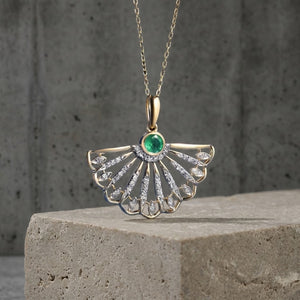 A gold fan-shaped pendant necklace with a central green emerald and clear stones, displayed on a textured stone block against a gray concrete wall.