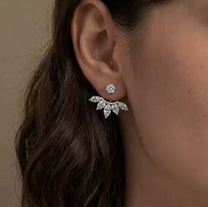 A close-up of a woman's ear wearing a silver-toned leaf-shaped stud earring with a small round stud above it, against a soft beige background.