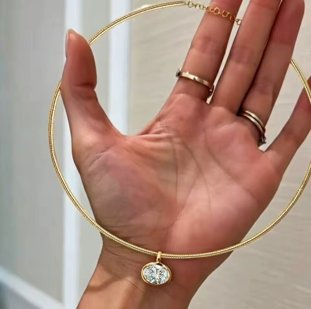 A gold snake chain necklace with an oval bezel-set clear gemstone pendant, shown worn around an open hand. The hand also has two gold rings on the fingers.