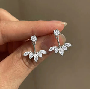 A close-up of a hand holding a pair of silver floral stud earrings with clear marquise-cut stones, against a soft beige background.