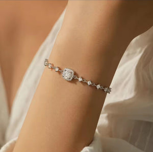 A delicate silver bracelet with a central cushion-cut diamond and smaller accent moissanite diamonds, shown worn on a person's wrist against a soft white fabric background.