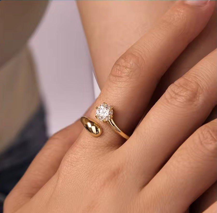 A close-up of a person's hand wearing a gold ring with a round clear gemstone, alongside another gold ring on the middle finger, set against a soft blurred background.