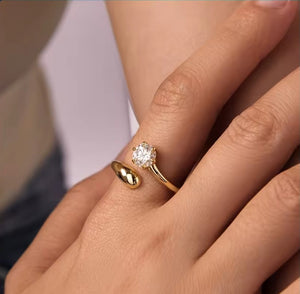 A close-up of a person's hand wearing a gold ring with a round clear gemstone, alongside another gold ring on the middle finger, set against a soft blurred background.