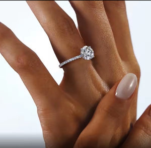 A close-up of a hand wearing a silver ring with a large round brilliant-cut centre moissanite stone and a pave band, set against a clean white background.
