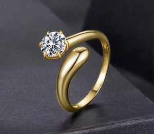 A gold solitaire ring with a round clear gemstone, displayed on a dark textured surface. The ring features a polished yellow gold band with a unique curved, open-ended design and a prong setting for the stone.