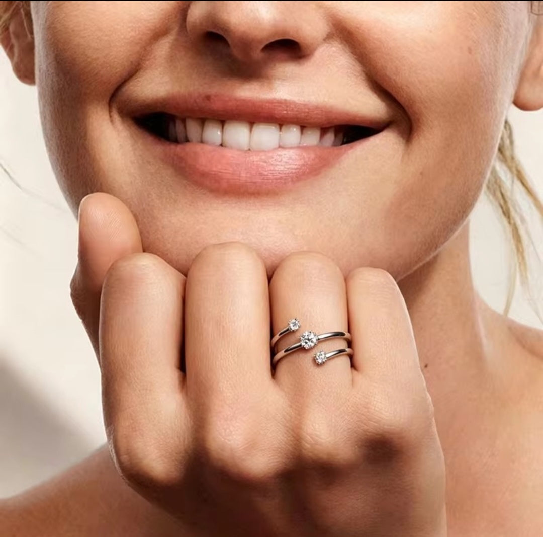 A close-up of a woman's hand and lower face wearing silver rings with small clear gemstones, set against a soft light beige background.
