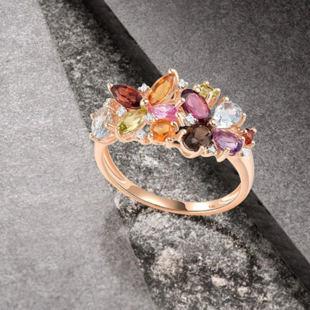 A multi-gemstone ring with a rose gold band, featuring a variety of colored gemstones set in a solid band, positioned on a textured grey background.