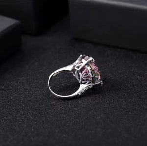 A silver ring with a large mystic quartz gemstone, photographed on a dark textured surface with black boxes in the background.