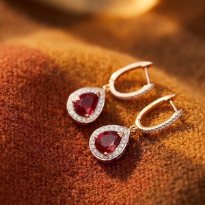 A pair of rose gold drop earrings with pear-shaped red ruby gemstones surrounded by small diamond clear stones, displayed on a textured brown surface.
