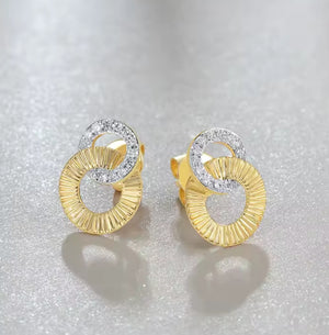 A pair of gold stud earrings with a textured, ribbed circular design and clear crystal natural diamond accents, displayed on a sparkly white surface.