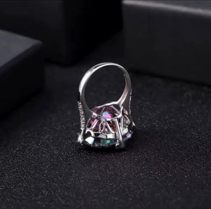 A silver ring with a large faceted iridescent natural quartz gemstone in a geometric setting, displayed on a textured black surface with dark boxes in the background.