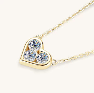 A gold heart-shaped pendant necklace featuring three round clear gemstones, shown on a delicate gold chain against a light cream background.