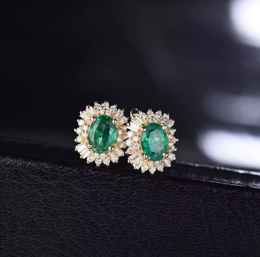 A pair of gold stud earrings with oval green emerald-cut centre stones surrounded by small clear diamond stones, displayed on a black textured surface against a dark blurred background.