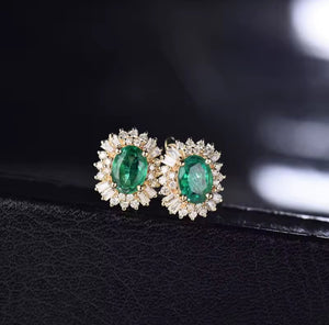 A pair of gold stud earrings with oval green emerald-cut centre stones surrounded by small clear diamond stones, displayed on a black textured surface against a dark blurred background.
