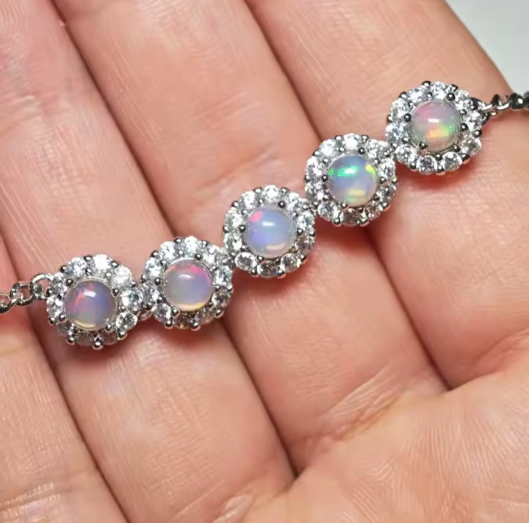 A close-up of a silver bracelet with five round iridescent opal-like stones, each surrounded by small clear stones, resting on a person's palm.