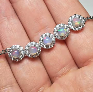A close-up of a silver bracelet with five round iridescent opal-like stones, each surrounded by small clear stones, resting on a person's palm.