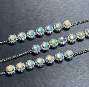 Three silver chain bracelets, each adorned with a row of round iridescent opal gemstones surrounded by small clear stones, displayed on a dark textured background.