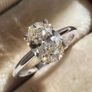 A silver solitaire ring with a large oval-cut clear gemstone, resting on a soft beige velvet ring box.