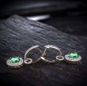 A pair of gold hoop earrings with oval green emerald gemstones surrounded by diamond stones, displayed on a dark textured stone surface.