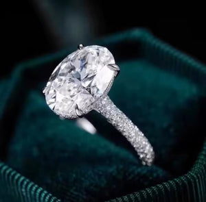 A close-up of a large oval-cut diamond ring with a pavé band, resting inside a dark green velvet ring box.