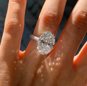 An oval-cut diamond ring with a silver-toned band, worn on a person's finger under bright sunlight.