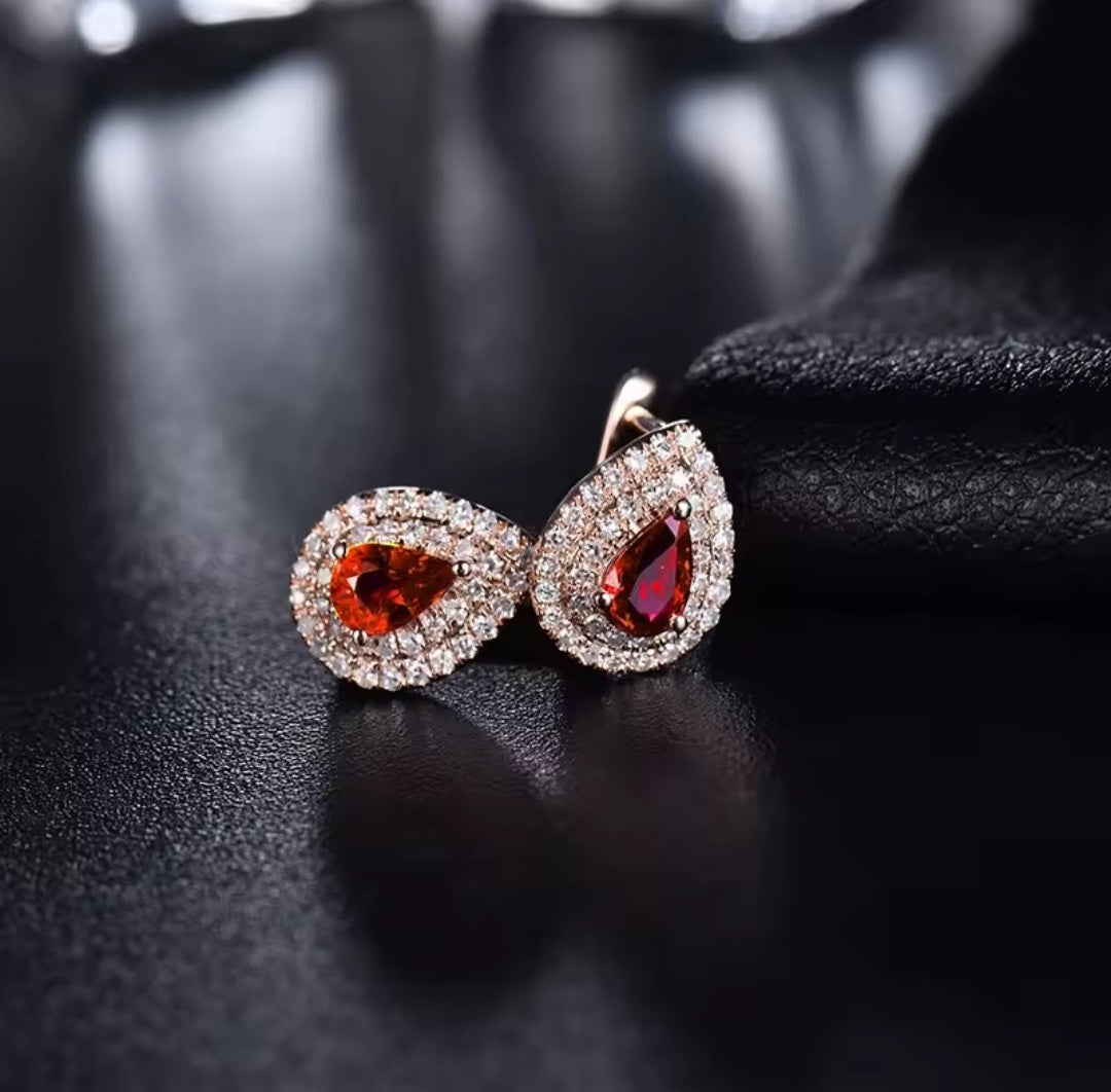 A pair of teardrop-shaped earrings with sparkling clear diamond halo settings and central deep red ruby gemstones, displayed on a dark textured surface.