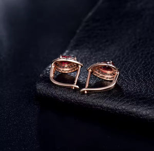 A pair of rose gold hoop earrings with oval red ruby gemstone centres and small clear natural diamond accent stones, displayed on a textured black leather surface.
