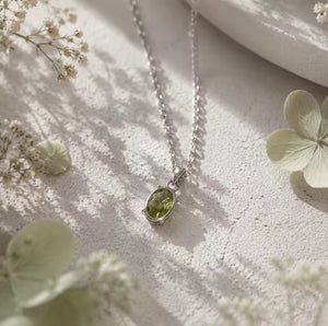 A silver pendant necklace with an oval green peridot gemstone, displayed on a textured light surface with soft dried flowers and shadows.