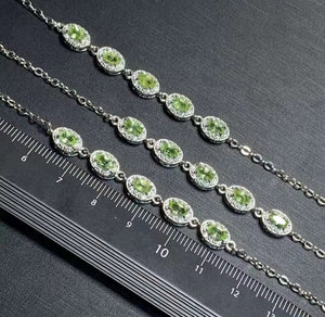 A silver-toned chain bracelet featuring multiple oval green gemstone links surrounded by small clear stones, shown alongside a black ruler for scale on a dark textured background.